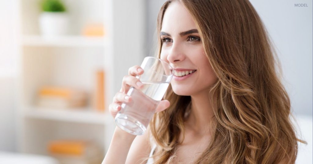 Close-up of woman with clear skin (model) standing in the kitchen drinking water.